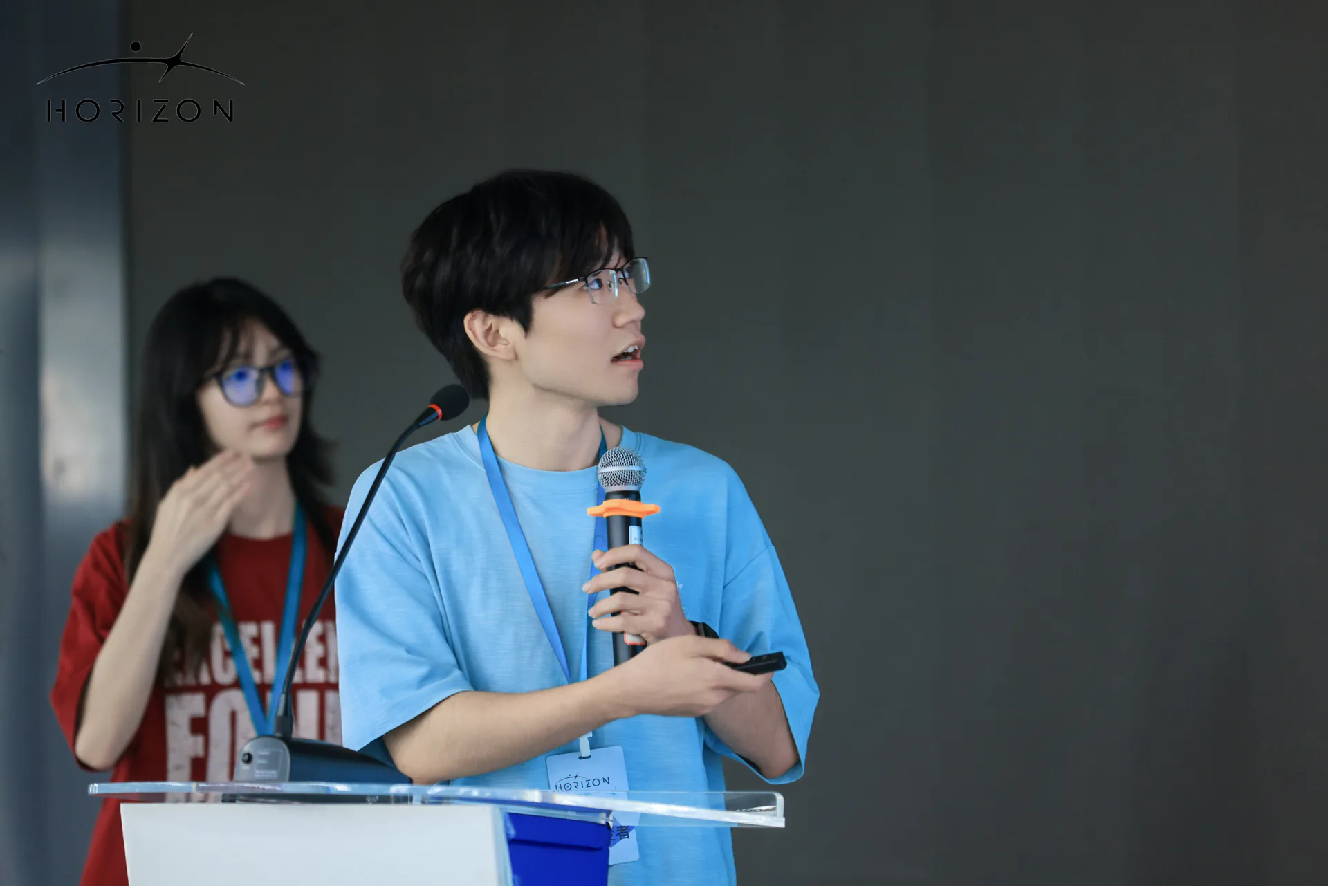 一名戴眼镜的年轻人在讲台前手持麦克风进行分享